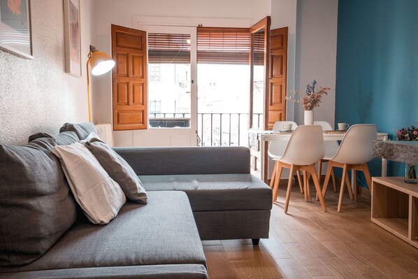 Ein gemütliches Wohnzimmer mit grauem Sofa, Holzboden und einem Essbereich mit weißen Stühlen und einem Tisch.