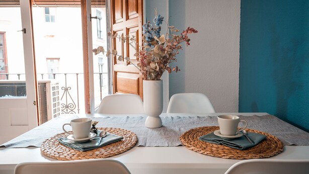A dining table with two white cups and saucers, a vase with dried flowers, and woven placemats.
