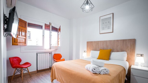 A hotel room with a large bed, a red chair, a wooden headboard, and a window with blinds. - QH Granada Center 13 (Granada)