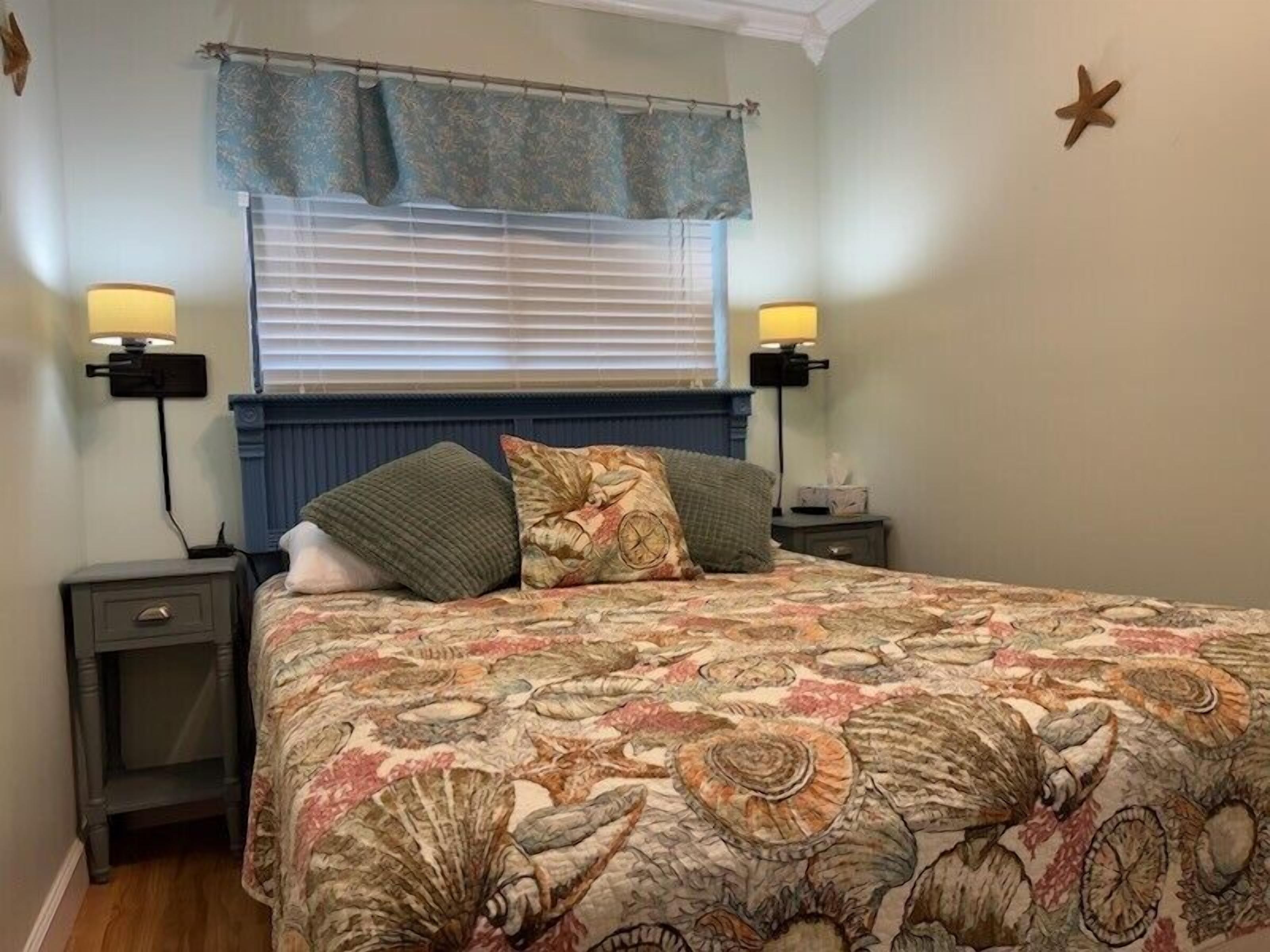 A bedroom with a bed, two bedside tables, a nightstand, a window with blinds, and a wall-mounted star decoration.