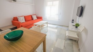 A modern living room with a wooden table, a red sofa, white chairs, and a TV mounted on the wall. - QH Aliatar Suites 01 (Granada)