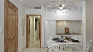 A modern kitchen with white cabinetry, a dining table set for four, and a unique pendant light. - Quality Homes Granada - QH San Juan de Dios (Granada)