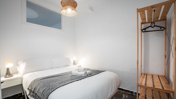 A bedroom with a bed, bedside lamps, a hanging light fixture, a wooden bench, and a wall-mounted coat rack. - Quality Homes Granada - QH Ayuntamiento 02 (Granada)