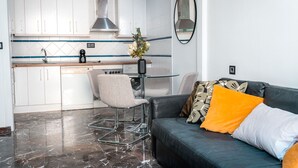 Ett kompakt bostadsutrymme med ett litet kök, en matplats med två stolar, en soffa med kuddar och ett marmorgolv.