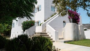 A white building with a staircase leading to the entrance, surrounded by greenery and flowers.