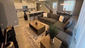 A modern living room with a large sectional sofa, a coffee table, and a dining area with a chandelier. - Magical 5BR w Pool Minutes From Disney 16161 (Clermont)