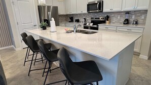 A modern kitchen with a large island, bar stools, stainless steel appliances, and pendant lighting. - Magical 5BR w Pool Minutes From Disney 16161 (Clermont)