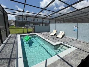 Une piscine avec un gros bouée en forme d’alligator vert, deux chaises longues et un panneau indiquant les règles de la piscine, fixé au mur.