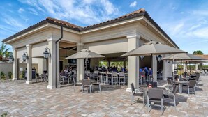 An outdoor patio area with multiple tables and chairs, covered by umbrellas, and a building with a tiled roof. - Golf Course Townhome Resort Amenities 2B Luxury (Bradenton)