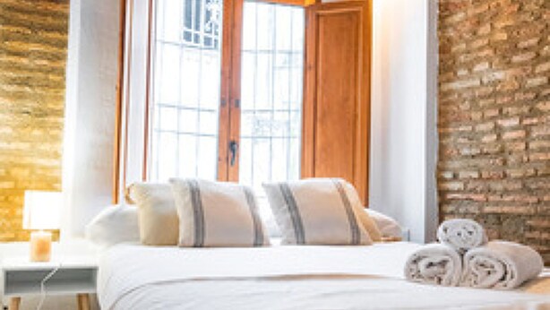 A bedroom with a large bed, wooden headboard, stone wall, and a window with wooden shutters. - Quality Homes Granada - QH Correo Viejo 04 (Granada)
