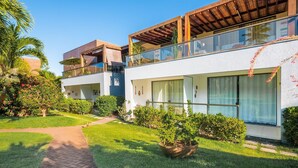 A modern two-story building with a glass balcony, surrounded by lush greenery and a paved pathway. - Condominium on the Coast in Imbassai - SOV (Mata de São João)