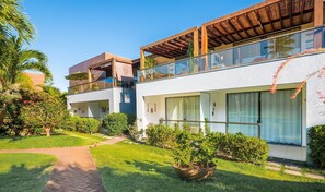A modern two-story building with a glass balcony, surrounded by lush greenery and a paved pathway. - Condominium on the Coast in Imbassai - SOV (Mata de São João)