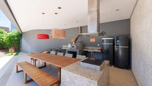 An outdoor kitchen area with a wooden table, benches, hanging pendant lights, a built-in oven, a stainless steel refrigerator, and a gas stove. - Condominium on the Coast in Imbassai - SOV (Mata de São João)