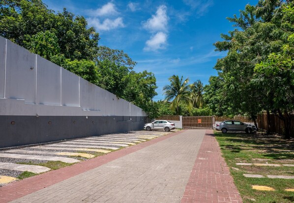 Un chemin pavé mène à une entrée fermée par un portail, le tout étant entouré d’une végétation luxuriante et surplombé par un ciel d’un bleu limpide.