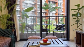 A balcony with a view of a multi-story building, a table with a bowl of fruit, and two patterned cushions. - Luxury Garden-View 1BHK I Near Candolim Beach I Free Parking I Fast Wifi (Candolim)