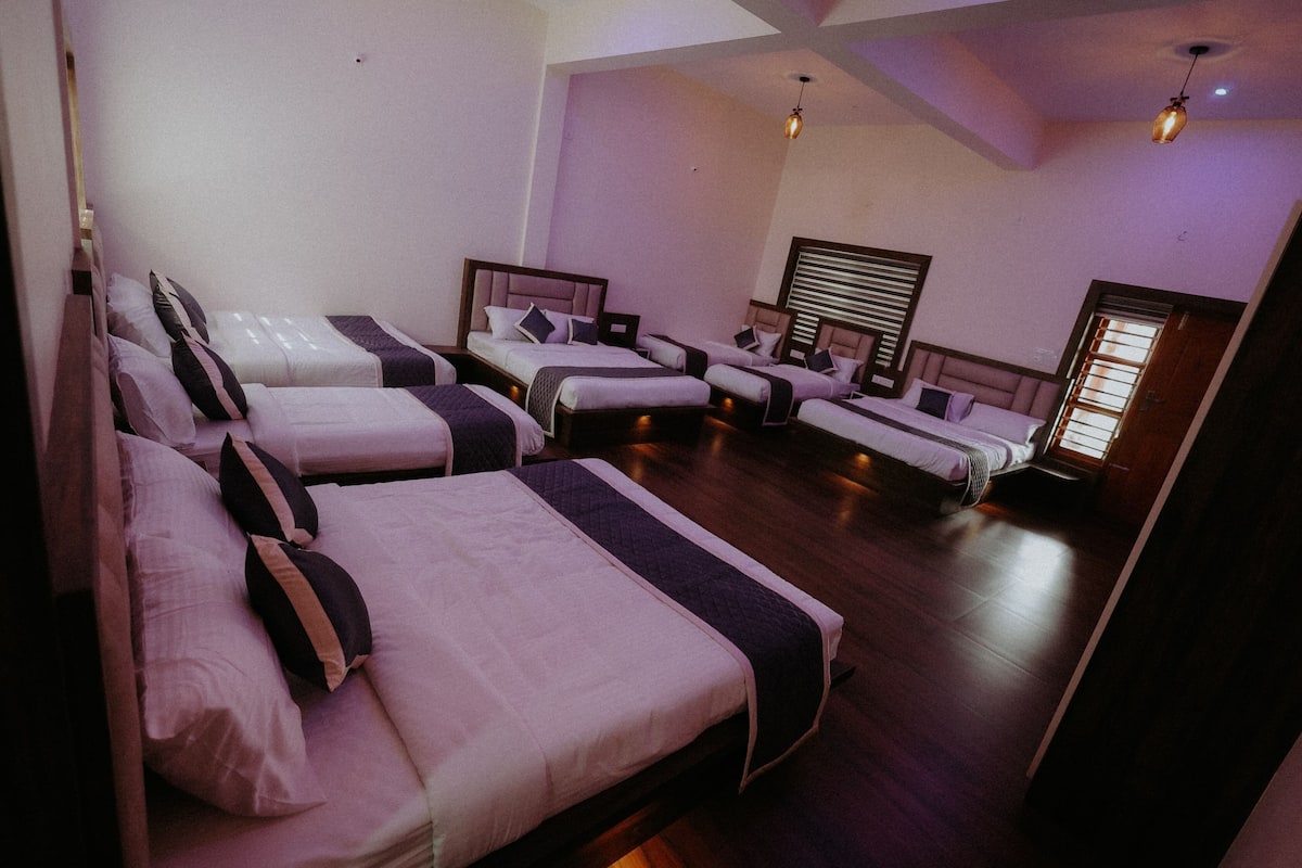 Une chambre d’hôtel avec quatre lits simples disposés en rangée, un plancher en bois et une grande fenêtre avec des stores.
