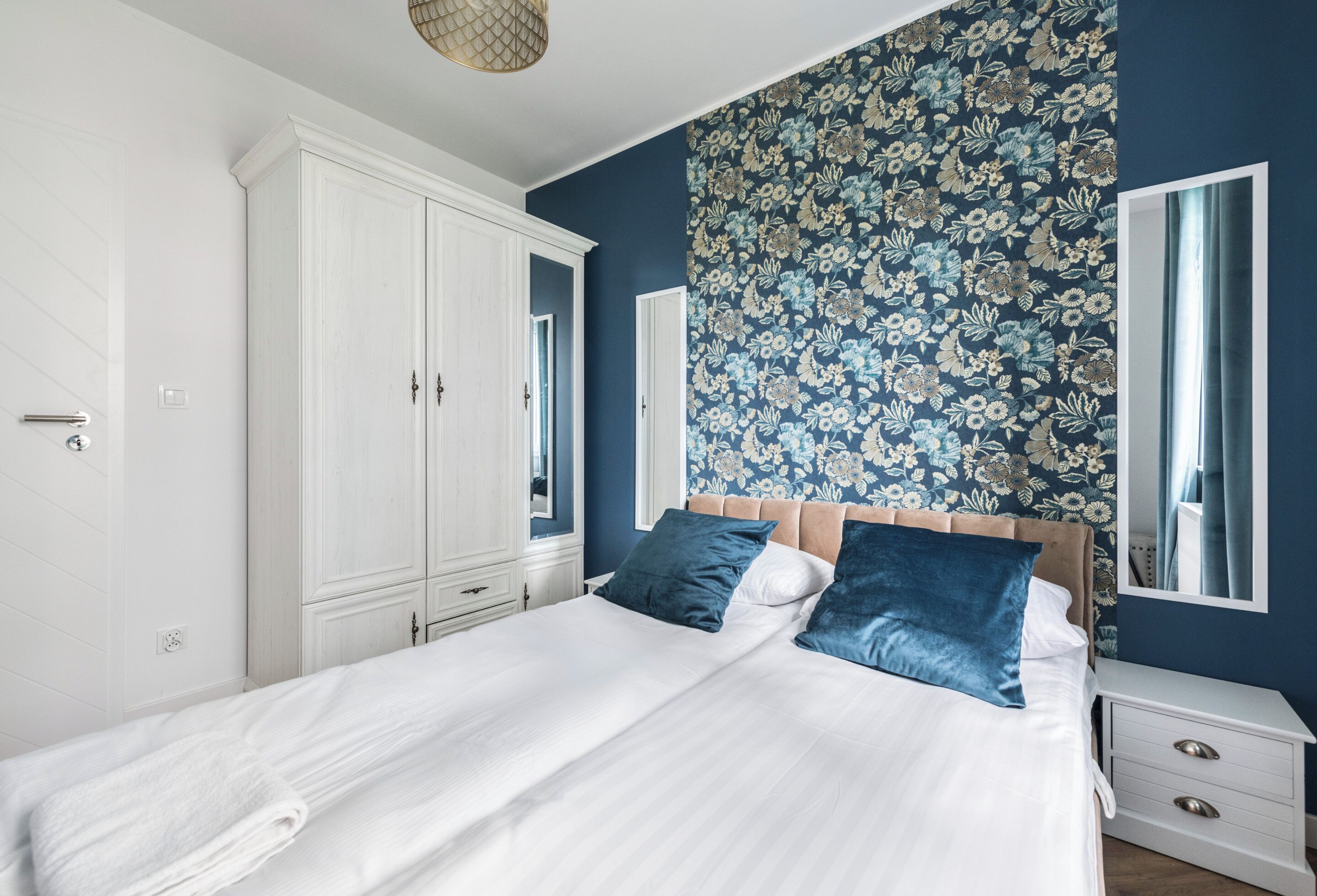 A bedroom with a large bed, white wardrobe, and a floral wallpaper.
