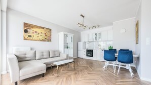A modern living room with a sofa, coffee table, and blue chairs. It features a kitchen with white cabinets and a dining area.