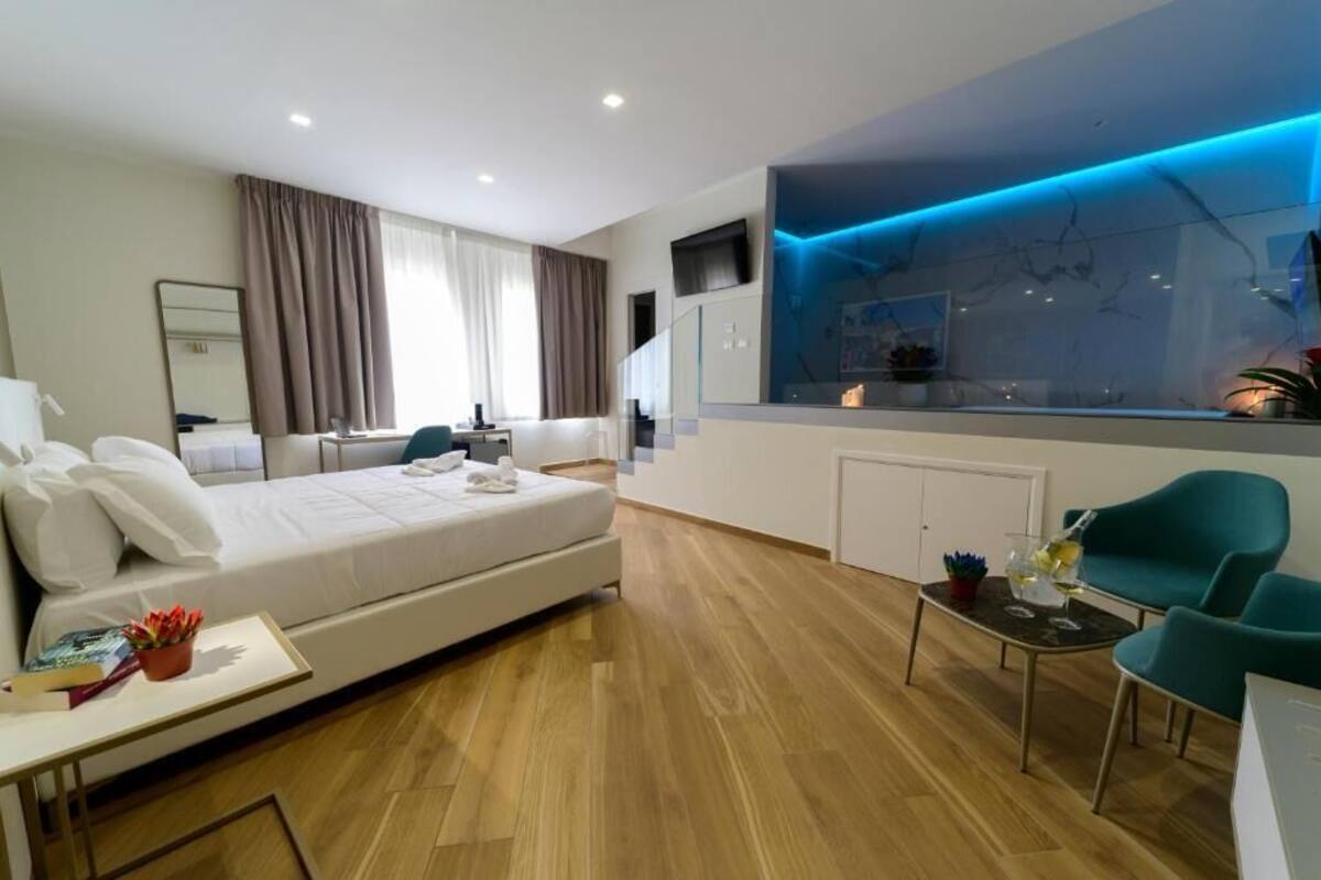 A modern hotel room with a large bed, a desk, a dining area, and a view of the city.