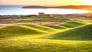 Un campo de golf con onduladas colinas verdes y vista al mar y al pueblo en el fondo.