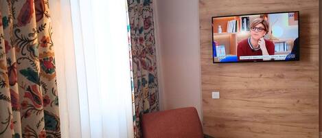 Habitación de hotel con una televisión instalada en una pared de madera, un sofá, dos sillones, una mesita redonda y una lámpara de pie.