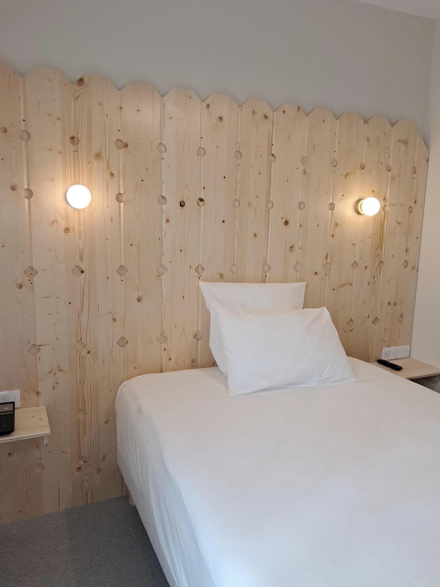 Un dormitorio con cabecera de madera, dos lámparas de noche, una cama con ropa de cama blanca y una mesita de noche con control remoto.