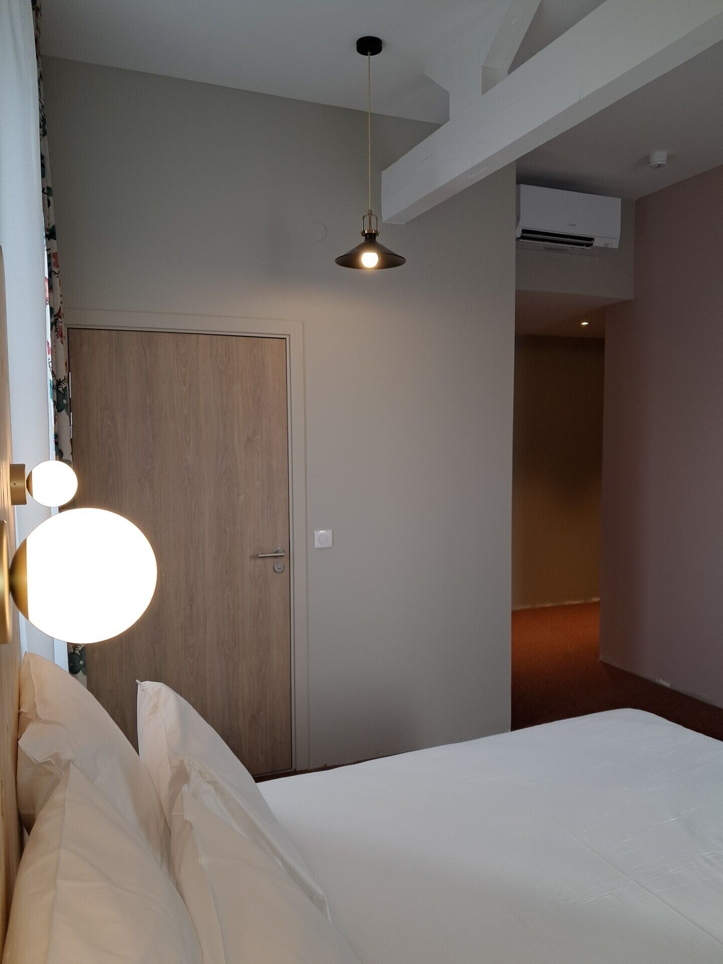 A hotel room with a bed, a door, a ceiling light, and an air conditioning unit.