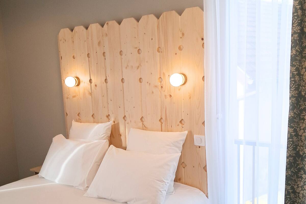A bedroom with a wooden headboard, two bedside lamps, a bed with white linens, and a window with sheer curtains.