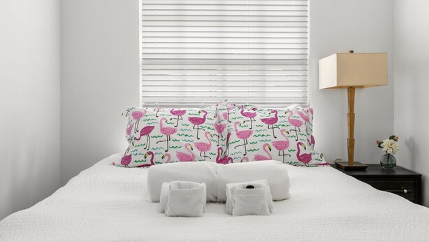 A neatly made bed with white bedding and pink flamingo patterned pillows, a bedside lamp, and a small vase with flowers on a nightstand.