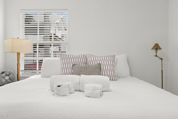 A neatly made bed with white linens, a bedside lamp, and a window with blinds.