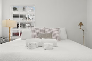 A neatly made bed with white linens and a striped pillow, a standing lamp, and a window with blinds.