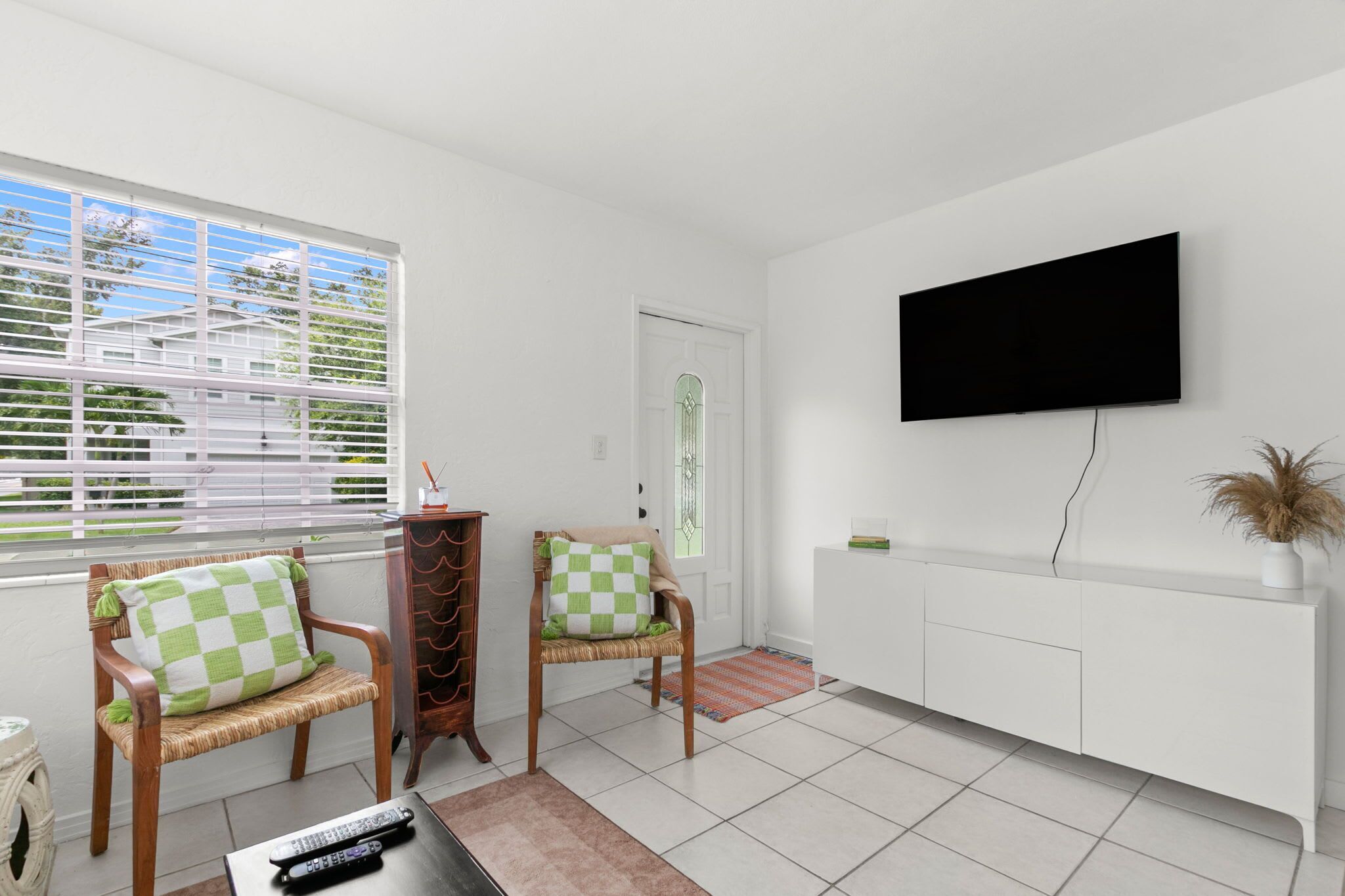 A living room with two wicker chairs, a television mounted on the wall, and a window with blinds.