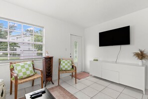 A living room with two wicker chairs, a television mounted on the wall, and a window with blinds.