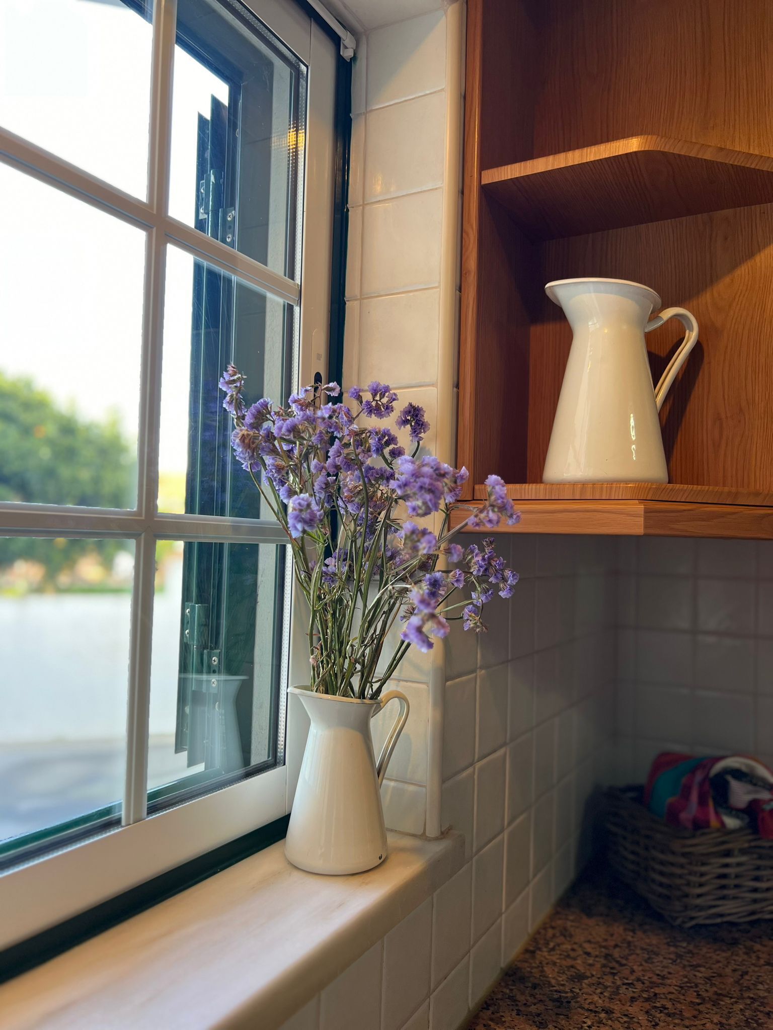 A kitchen with a window, a white pitcher, and a vase with purple flowers.
