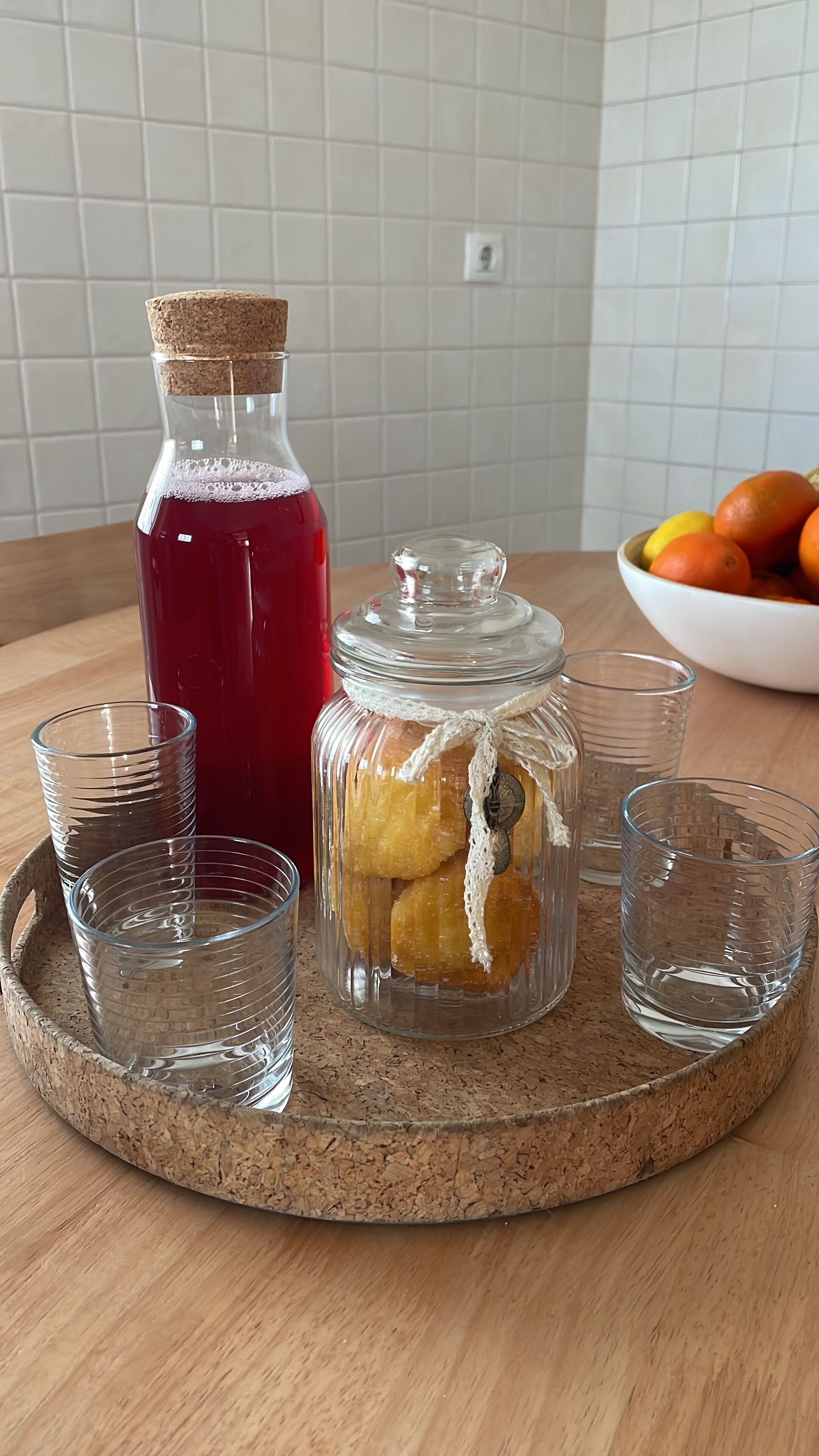 A kitchen counter with a corked glass bottle, a glass jar with citrus peels, and two empty glasses on a cork coaster.
