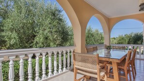 Een overdekte eethoek met een glazen tafel en rieten stoelen, omgeven door een witte balustrade en groen.