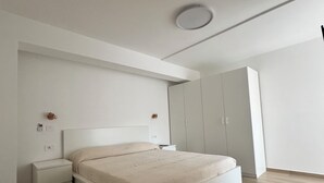 A modern bedroom with a large bed, a nightstand, a wardrobe, and a ceiling light. - Good House Holidays - THREE ROOM (Barano D'ischia)