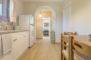 Eine Küche mit weißen Schränken, einem Holztisch und Stühlen. Es gibt einen Kühlschrank, eine Spüle und ein Fenster mit Vorhang.