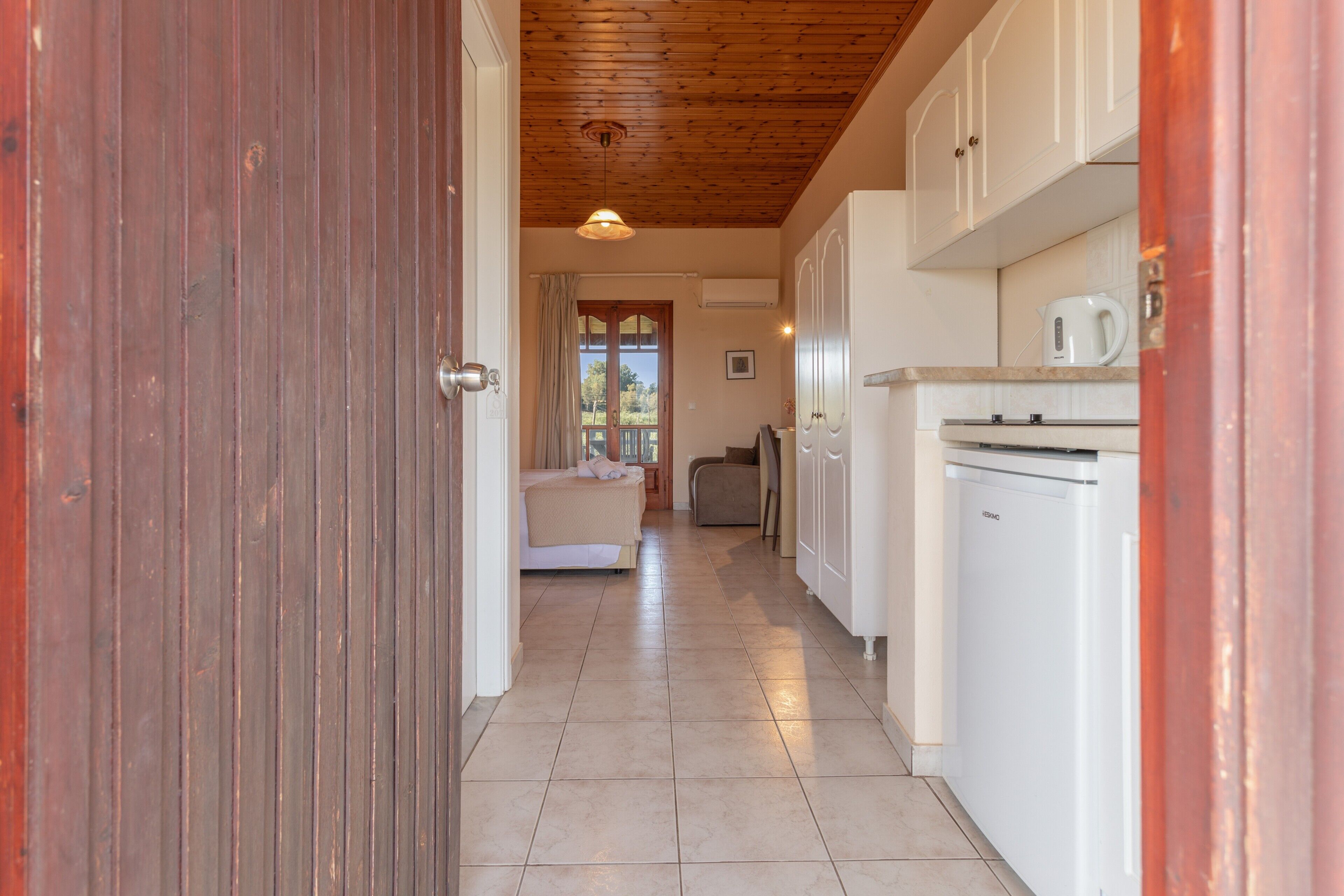 A hallway leading to a bedroom with a wooden ceiling, a kitchen with white cabinets, and a view through a window.