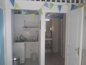 Een compacte keuken met een spoelbak, een kleine koelkast en een prullenbak. Een deur leidt naar een badkamer met een spiegel en een wastafel.