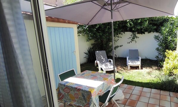 Een terras buiten met een tafel, stoelen, een parasol en een blauwe deur.