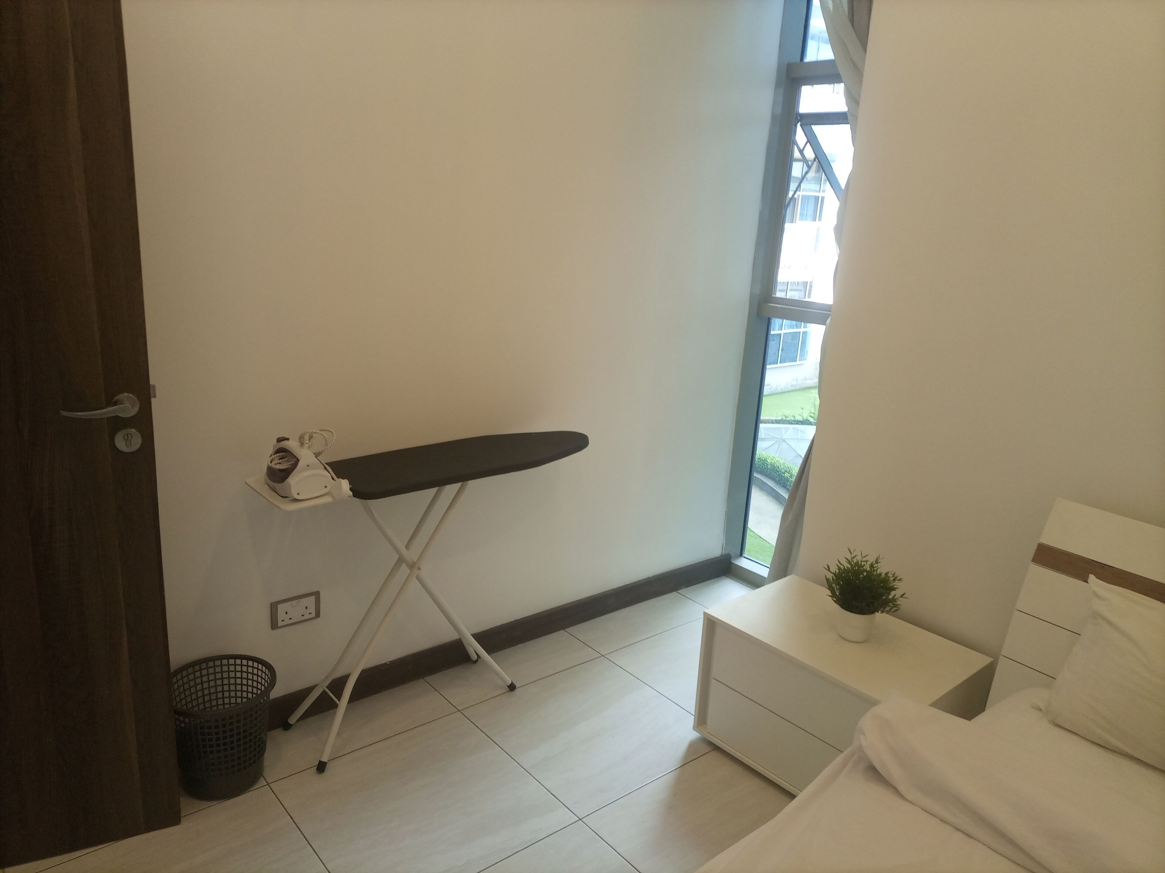 Une chambre avec un lit, une petite table de chevet ornée d’une plante, une fenêtre donnant sur un paysage verdoyant et une planche à repasser adossée au mur.