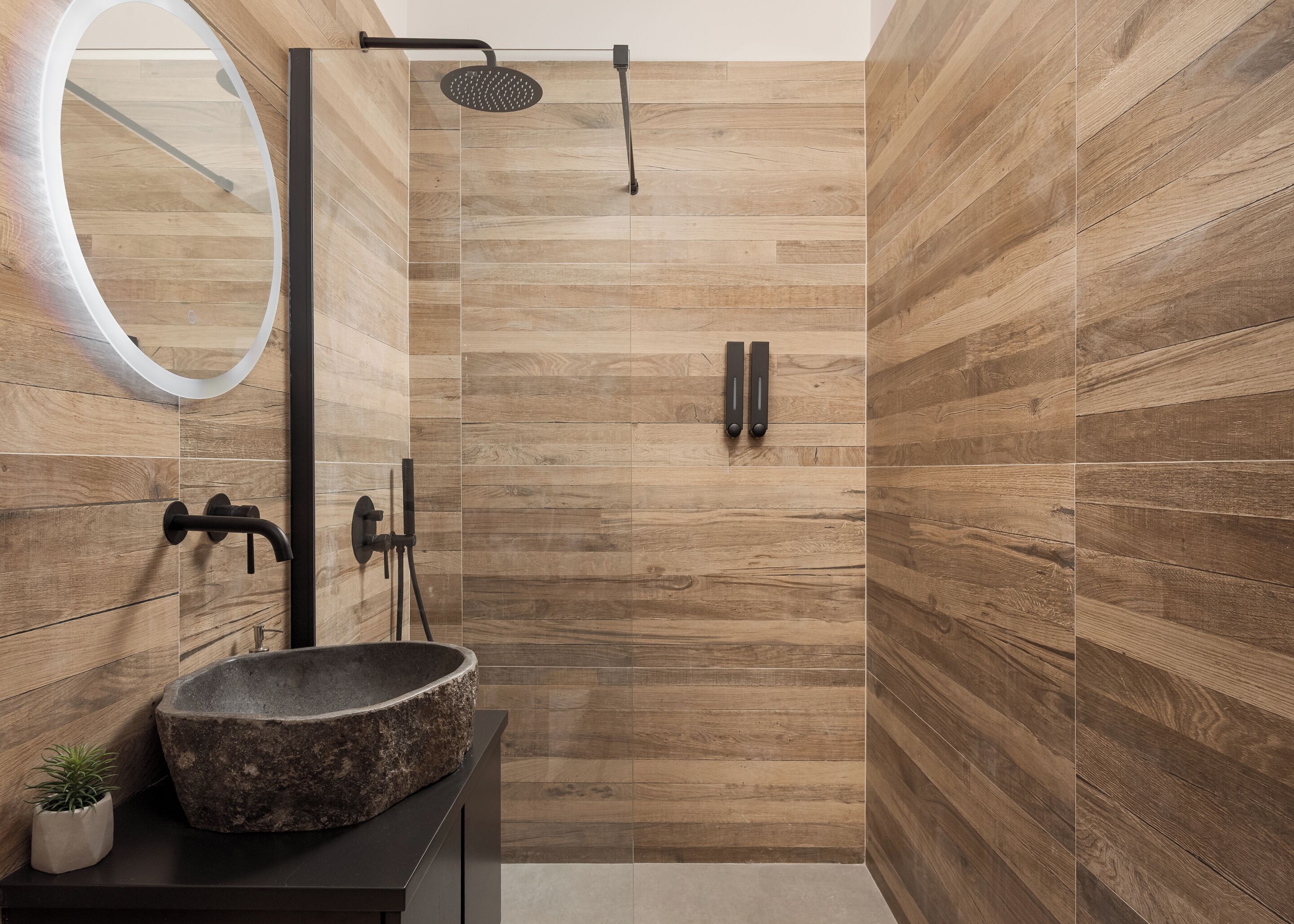 Un baño moderno con pared de madera, un espejo redondo, lavamanos de piedra y ducha con cabezal de ducha manual.