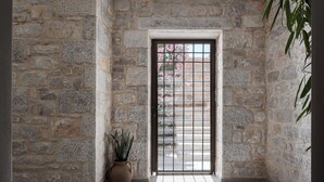 Un couloir aux murs de pierre, avec une grille métallique, un pot de fleurs et un sol carrelé.