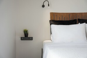 A neatly made bed with a headboard, a small shelf with a plant, and a wall-mounted light fixture.
