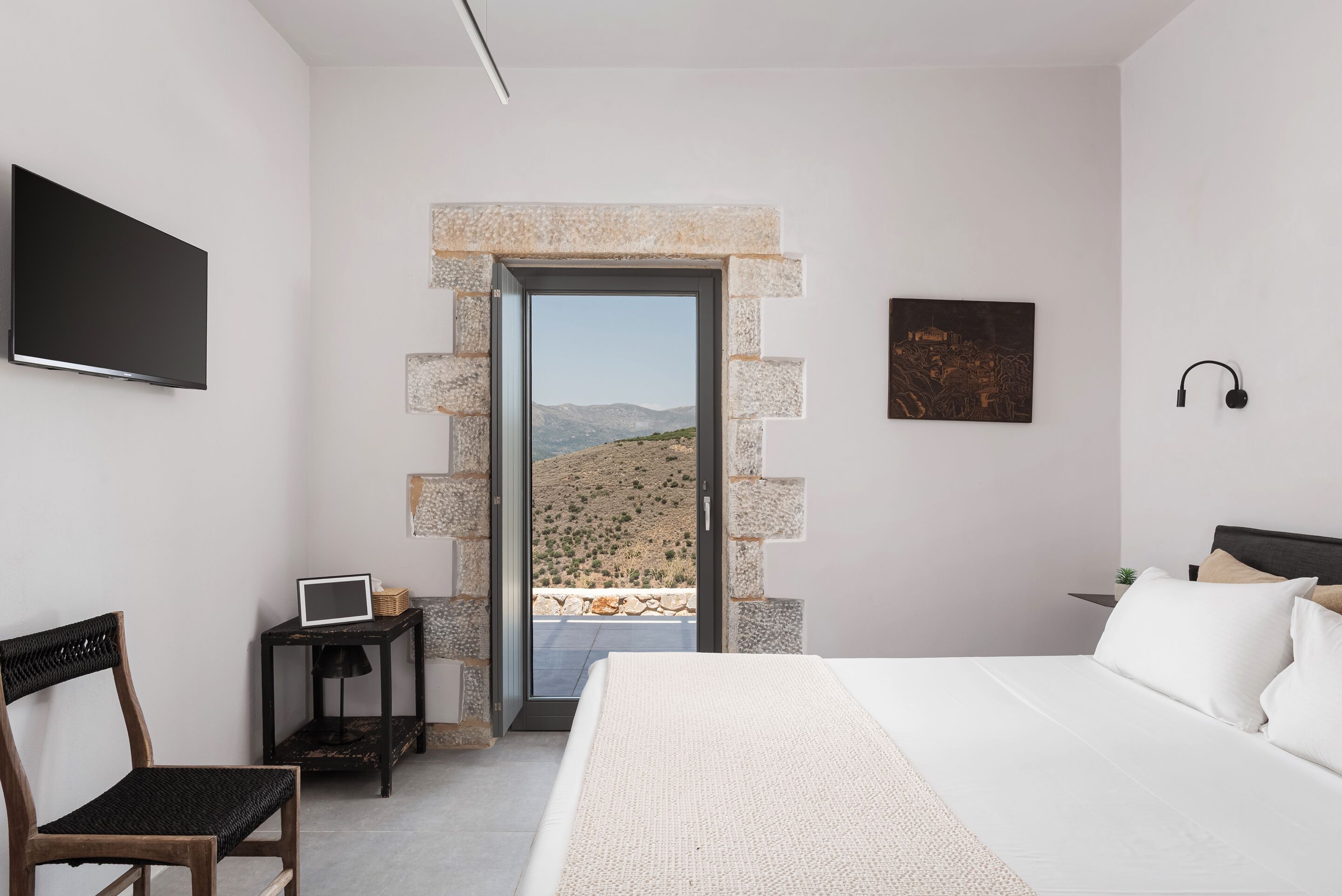 Un dormitorio con cama, escritorio, silla, televisor y puerta que da a un balcón con vistas al paisaje.