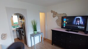 A living room with a television displaying a show, a dining area with a table and chairs, and a kitchen visible through an archway.