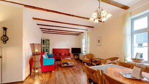A living room with a red sofa, a wooden coffee table, wicker chairs, a dining table with placemats, a chandelier, and a large window with curtains. - Apartment 'Kavaliershäuschen' with Mountain View, Shared Garden and Wi-Fi (Maring-Noviand)