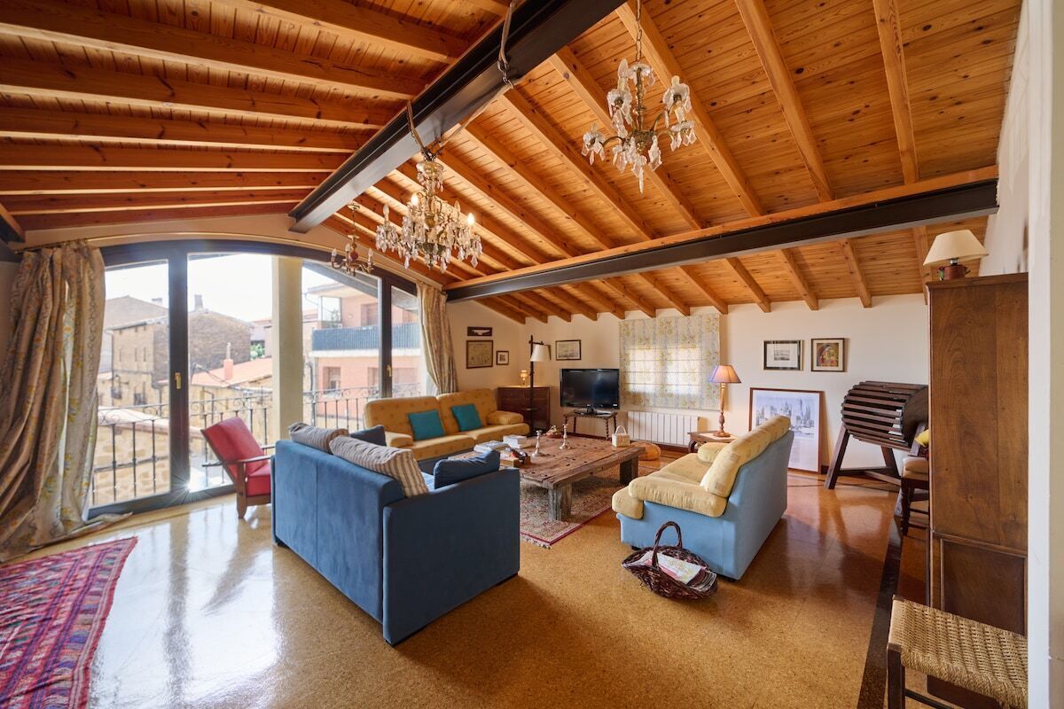 A spacious living room with a high wooden ceiling, a large window, and a balcony.