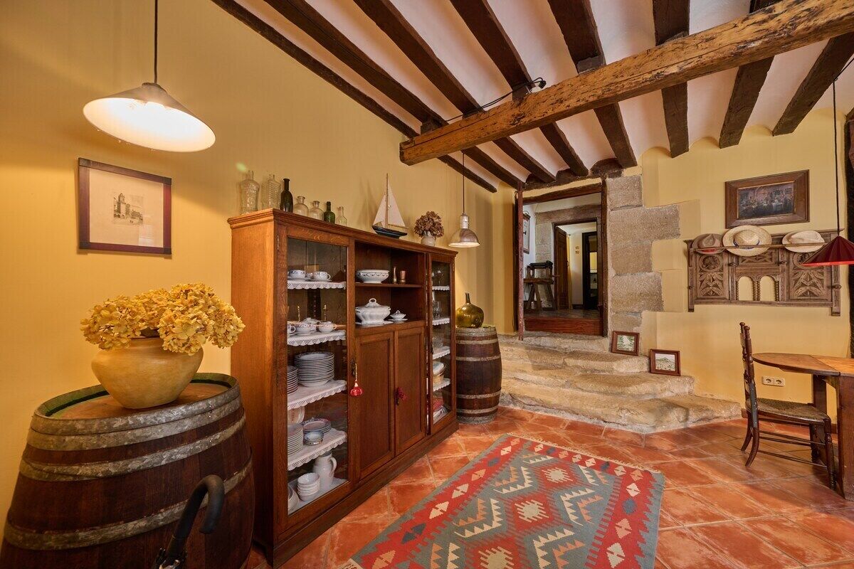 Een rustiek interieur met zichtbare houten balken, een houten kast met servies, een vloerkleed met patroon en een vat.